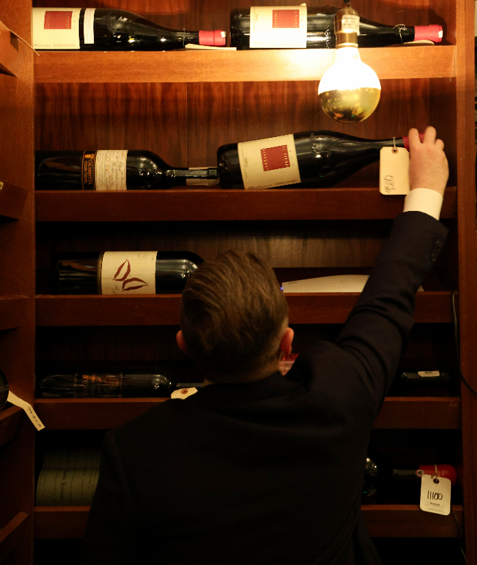 A person in a dark jacket reaches up toward a bottle on a warmly lit wooden wine shelf lined with large wine bottles and a hanging exposed light bulb.