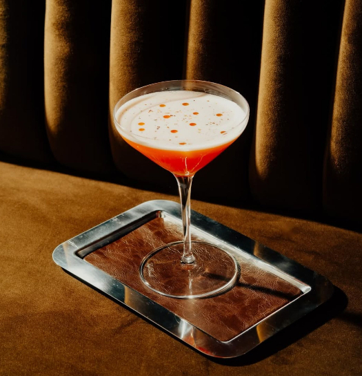 A pink-orange cocktail with a frothy top sits in a coupe glass on a small silver tray atop a brown upholstered bench, lit dramatically against a dark velvet backrest.
