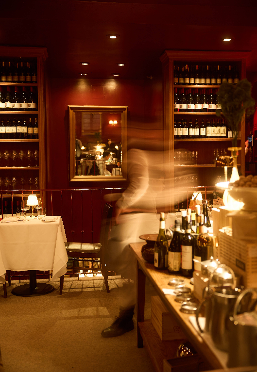 A warmly lit restaurant interior with walls of wine bottles and white tablecloth tables, while a blurred server moves through the room in the foreground.