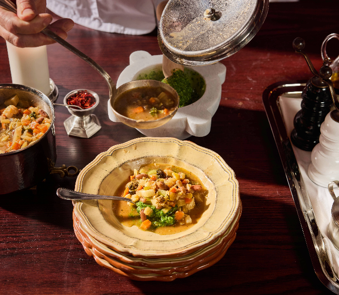 A bowl of minestrone sits on a dark wooden table, topped with diced vegetables and herbs, with a second serving dish and small condiments in the background.