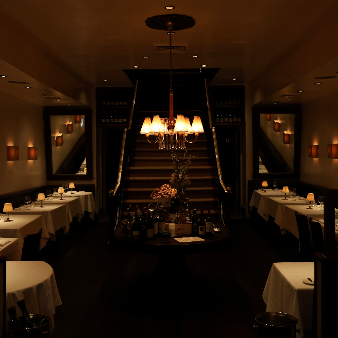 A dim, elegant restaurant dining room with white tablecloth tables arranged symmetrically around a central staircase, lit by a warm chandelier and small wall sconces.