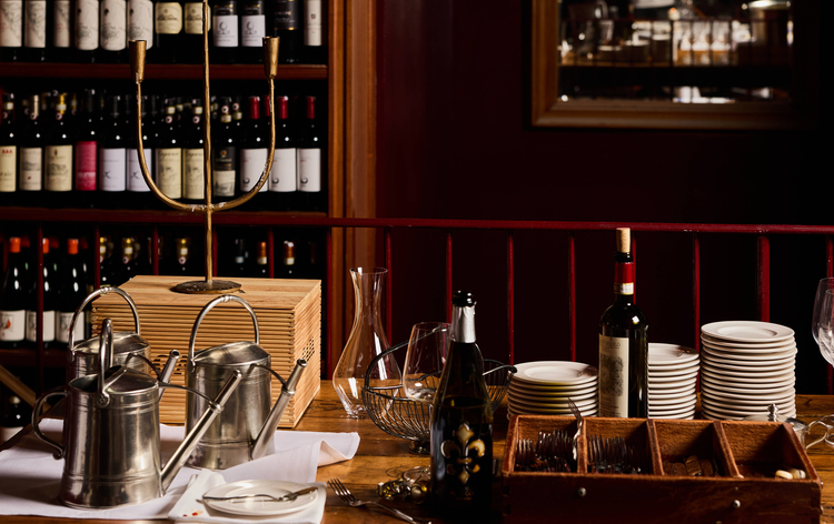 A wine-tasting setup with bottles, glasses, silver carafes, plates, and a wooden tray in a dimly lit wine shop.