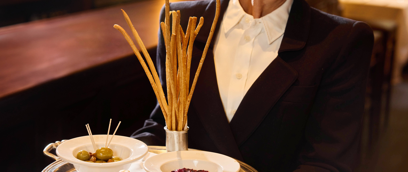 Waitress holding a tray with breadsticks, olives, and dried fruit in a restaurant setting.