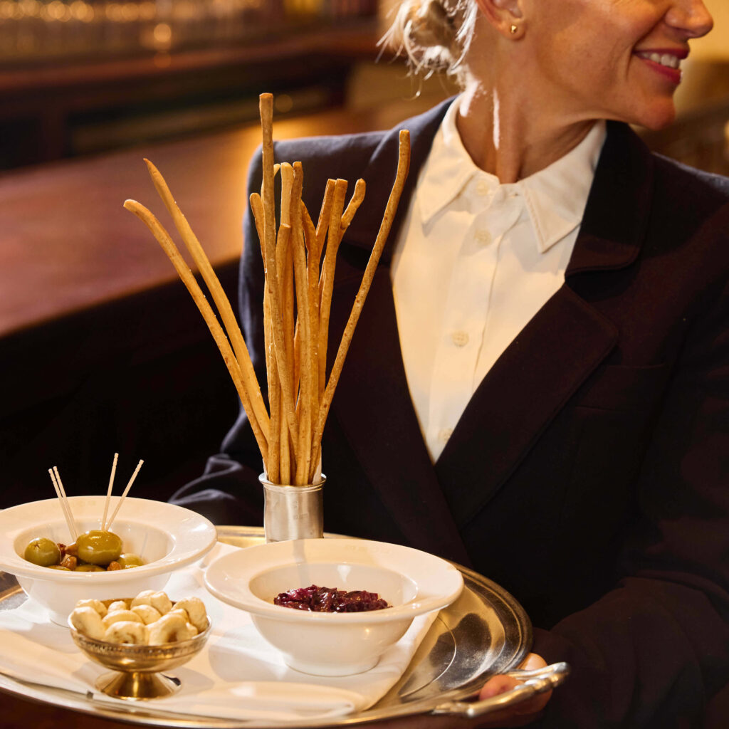 Server with Blue Suit holding a silver tray of antipasti small plates of olives, sun-dried tomatoes and marinated mushrooms.